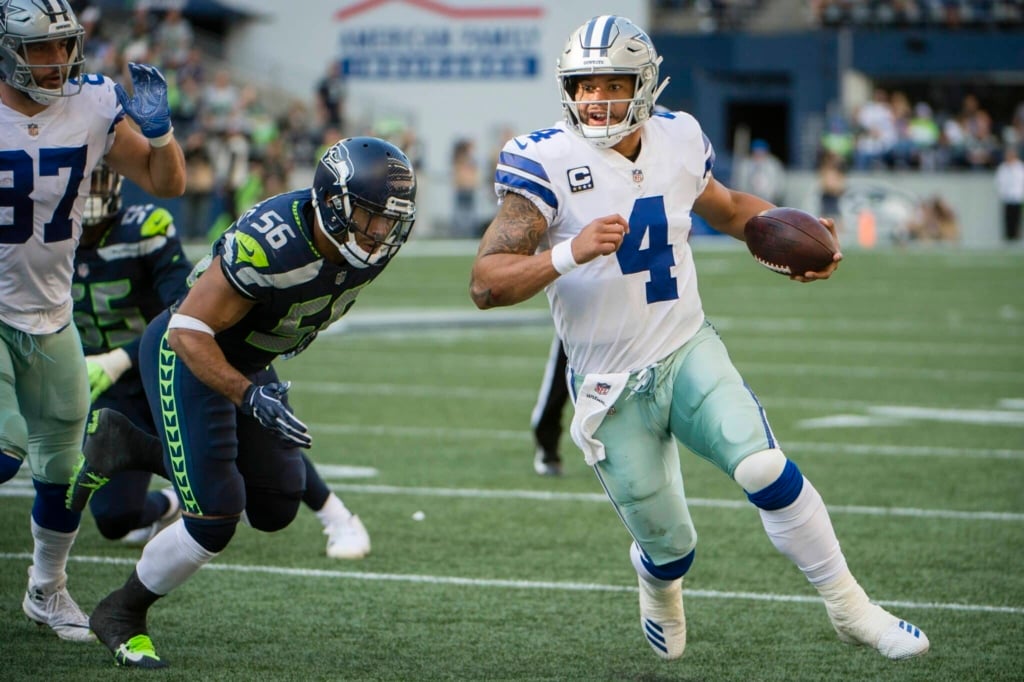 A football player in a white and blue uniform runs with the ball while being chased by an opponent in a blue and green uniform.