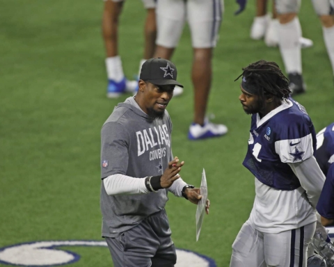 Dallas Cowboys coach in a grey outfit talks to player Trevon Diggs on the field with others in the background.