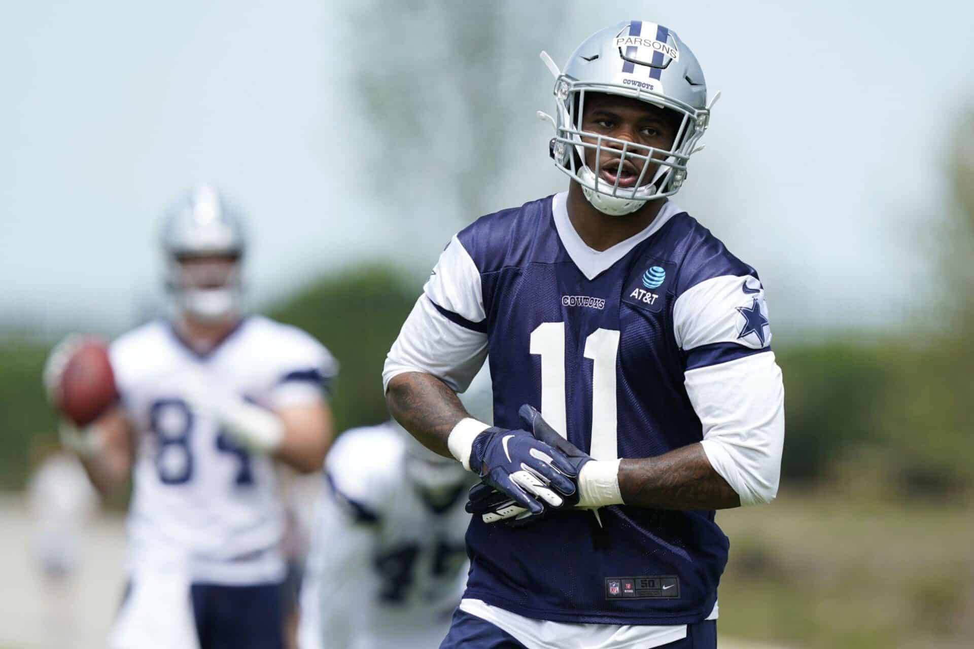 A football player in a navy and white uniform practices at Training Camp, with a teammate holding a football in the background.