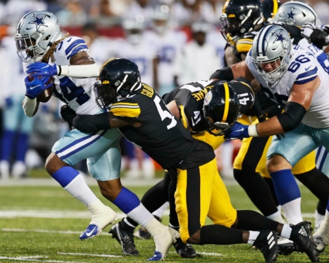 Football players from opposing teams in action on the field, one player carrying the ball while others attempt a tackle.