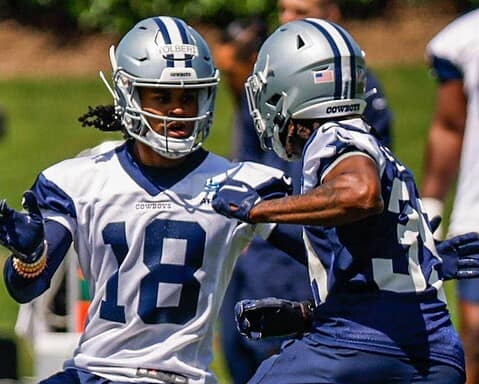 Two football players in Dallas Cowboys uniforms and helmets, including Jalen Tolbert, practice on the field with teammates in the background.