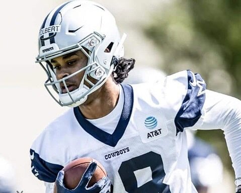 Football player in a white and blue uniform running with the ball during practice, wearing a helmet bearing the name "Jalen Tolbert," representing the Cowboys.