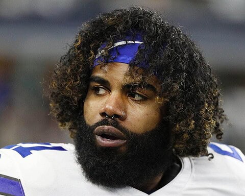 A football player with a beard and curly hair, resembling Ezekiel Elliott, wearing a white and blue jersey glances to the side on the field during preseason play.