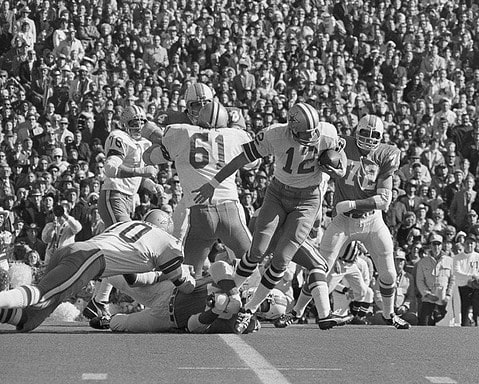 Black and white photo of a football game with players from both teams in action, featuring the Cowboys' legendary Roger Staubach. A crowd of spectators can be seen in the background.