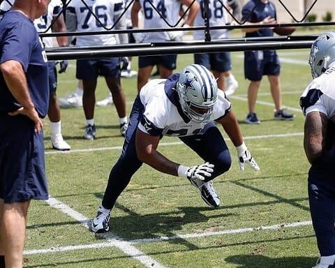 Football players in jerseys and helmets practice on a grassy field, with coaches observing them closely. Among them, Chuma Edoga stands out as he hones his skills with precision and determination.