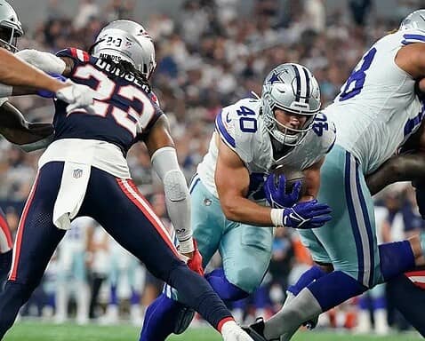 Football players from opposing teams in action during a game, with one team in blue and white uniforms and the other in blue and red. Hunter Luepke of the Cowboys makes a pivotal play that has fans on their feet.
