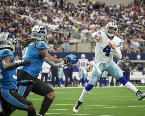 A football player in a white uniform, resembling Dak Prescott, throws a pass while three Panthers players in blue and black attempt to block the throw.
