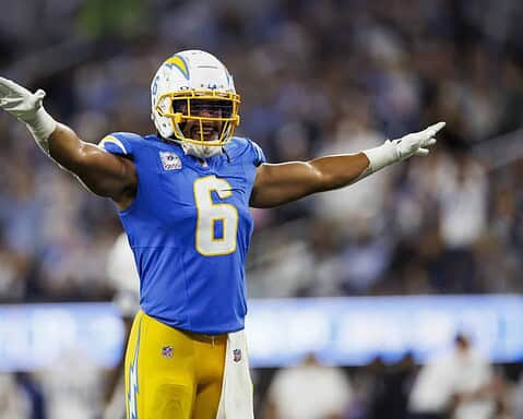 Football player in blue uniform with number 6 spreads arms wide on the field during a game. Crowd blurred in background.