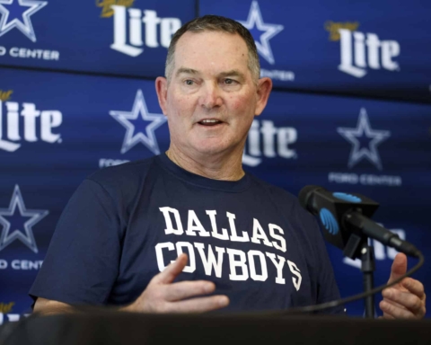A man wearing a Dallas Cowboys shirt speaks at a press conference with a backdrop of Cowboys logos and Miller Lite branding.