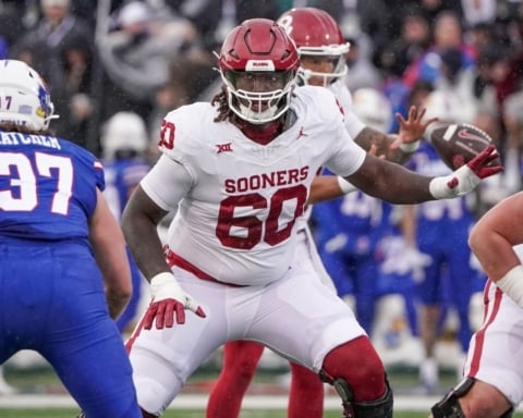 Tyler Guyton, a football player in an Oklahoma Sooners uniform, prepares to block an opponent from the opposing team wearing a blue uniform.
