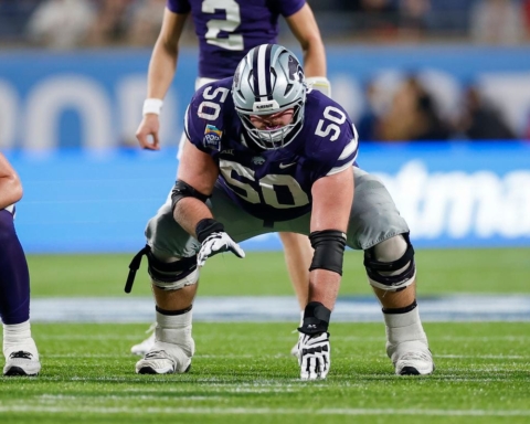 Football player in a crouched stance on the field, wearing a purple and gray uniform with the number 50. Cooper Beebe prepares to launch into action, determination evident in his eyes.