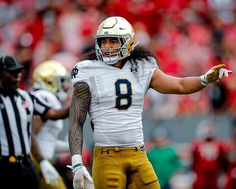 A football player in a white and yellow uniform with number 8, resembling Marist Liufau, stands on the field during a game, gesturing with his left hand.