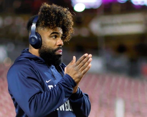 A man with headphones and curly hair, resembling Ezekiel Elliott in his focus, wears a dark blue hoodie as he stands with hands clasped in an outdoor setting.