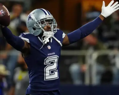 Football player in a blue jersey and helmet raises arms while holding a football on the field.