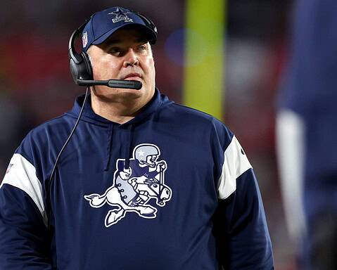 A dedicated coach in a navy sports hoodie and headset attentively observes the game, strategizing every move.