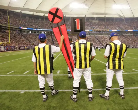 Three officials in striped uniforms hold yardage markers on the sidelines during a football game in a stadium, seamlessly integrating technology to ensure precise measurements and smooth gameplay.