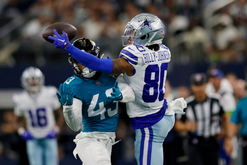 A football player in a blue uniform attempts to catch a pass, while Tyron Billy-Johnson, in a white and blue uniform, tries to block him.