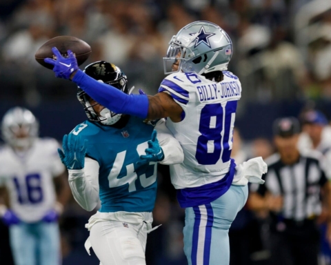 A football player in a blue uniform attempts to catch a pass, while Tyron Billy-Johnson, in a white and blue uniform, tries to block him.
