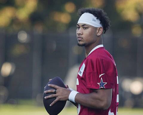 A football player in a red jersey holds a football, standing outdoors with a focused expression.