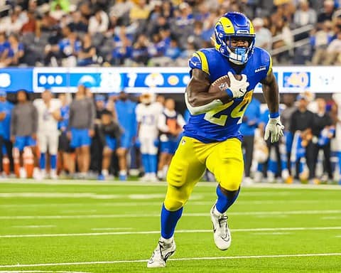 During Cowboys Training Camp, an RB in blue and yellow uniform runs with the ball during a game on a field with a crowd in the background, earning praise for his agility and performance.