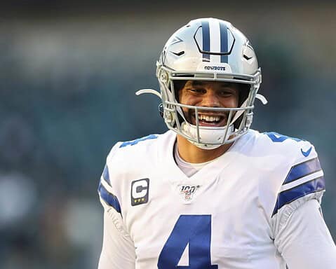 A football player in a white Dallas Cowboys jersey and helmet smiles on the field.