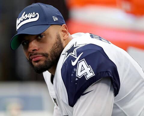 A football player in a Cowboys uniform and cap looks towards the camera while sitting on the sideline.