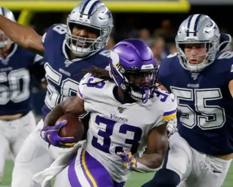 Football player in a purple jersey runs with the ball, pursued by three players in navy blue jerseys on a field.