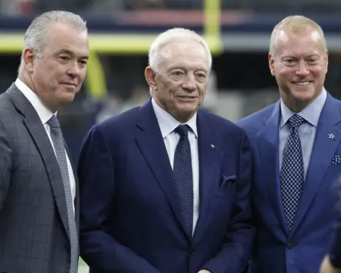 Three men in suits stand together, smiling, in a stadium setting.