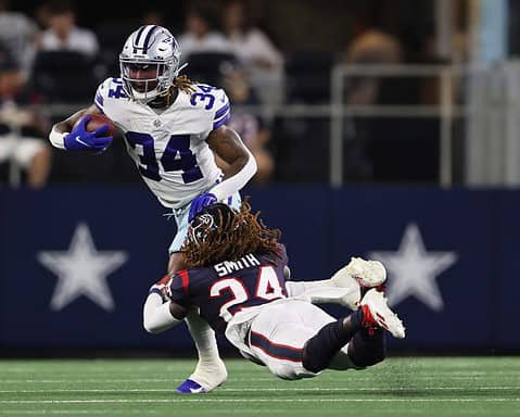 Football player in a white jersey runs with the ball while a player in a black jersey dives to tackle him.
