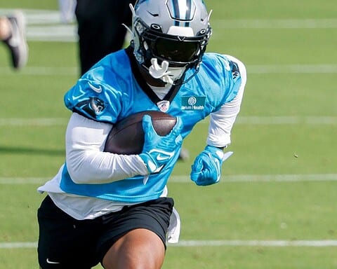 Football player in a blue jersey, holding a football, running on a grassy field during practice.