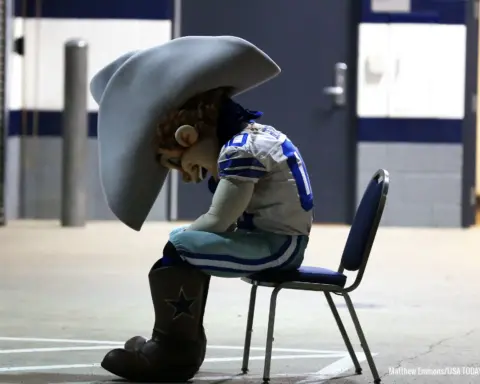 A sports mascot in cowboy attire sits on a chair with a slumped posture in a hallway.