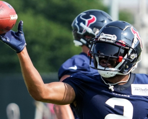 Football player in navy uniform practices throwing a football during a training session, with teammates in the background.