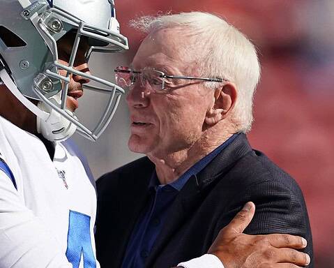 A football player in uniform and a man in a suit embrace on the field.