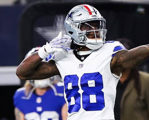 Football player in a white and blue uniform pointing while celebrating during a game. Fans and staff in the background.