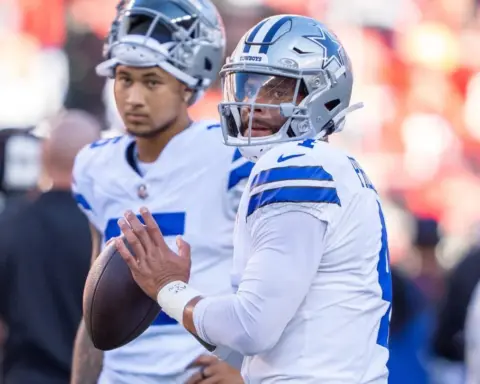 Two football players in white and blue uniforms, one holding a football, stand on the sideline during a game.