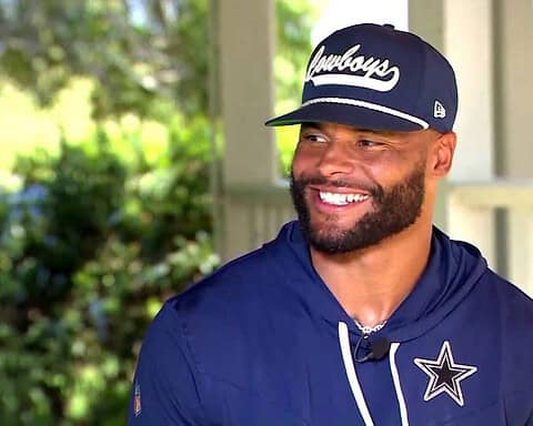 A man in a navy blue Cowboys cap and hoodie with a logo smiles while outdoors under a covered area.