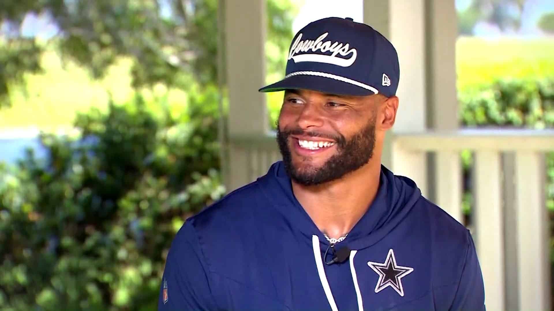 A man in a navy blue Cowboys cap and hoodie with a logo smiles while outdoors under a covered area.