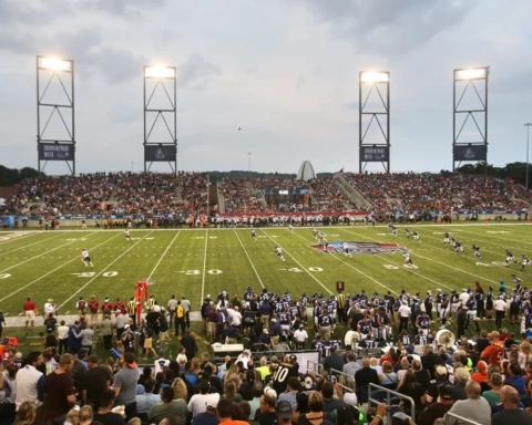 Packed football stadium with fans watching a game under bright lights during twilight, with the field in active play and incorporating recent changes to enhance the experience.