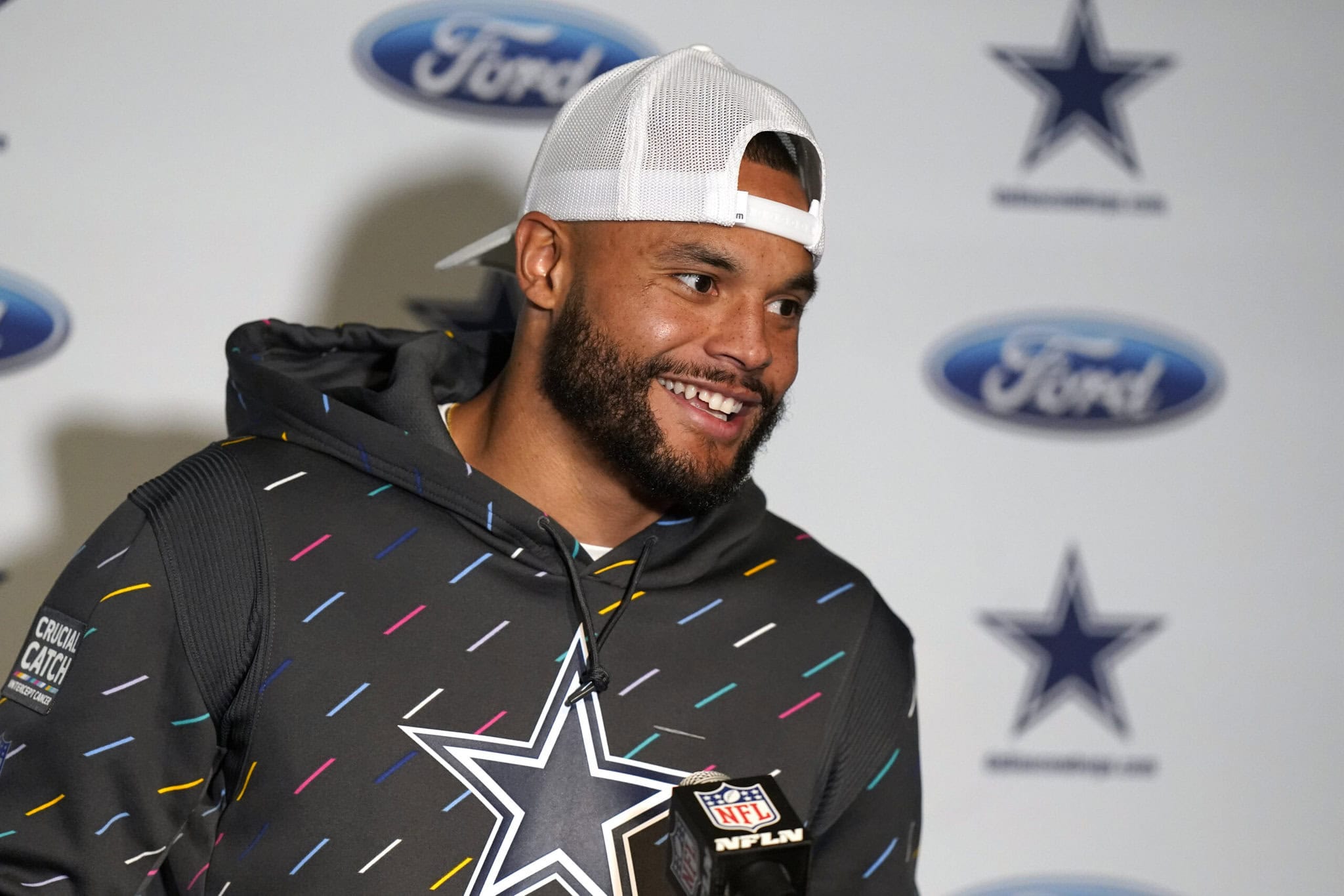A man in a hoodie and cap stands in front of a backdrop with Ford and Dallas Cowboys logos, smiling.