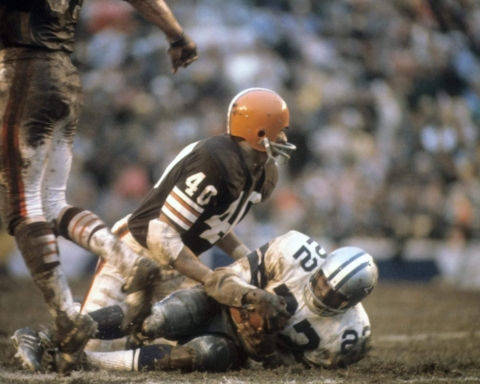 Two American football players in a tackle, one from a brown team and the other from a white and blue team, on a muddy field.