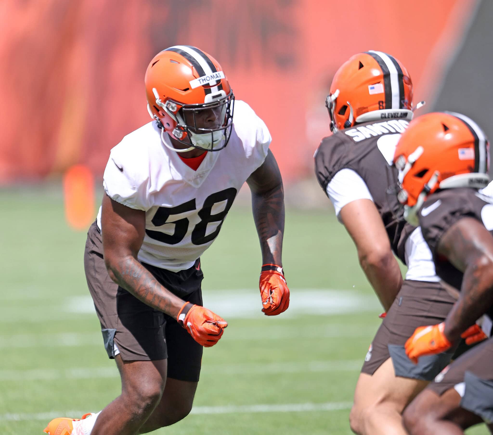 Football players in orange helmets and jerseys during practice on a green field. One player is in a defensive stance.