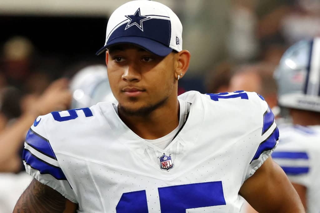 A football player wearing a Dallas Cowboys uniform and cap stands on the sidelines during a game.