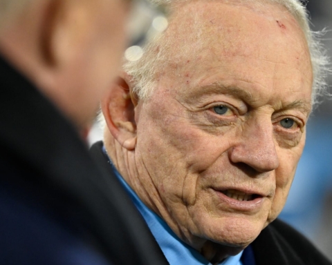 An elderly man with thinning white hair and a light blue shirt talks to another person, who is partially visible.