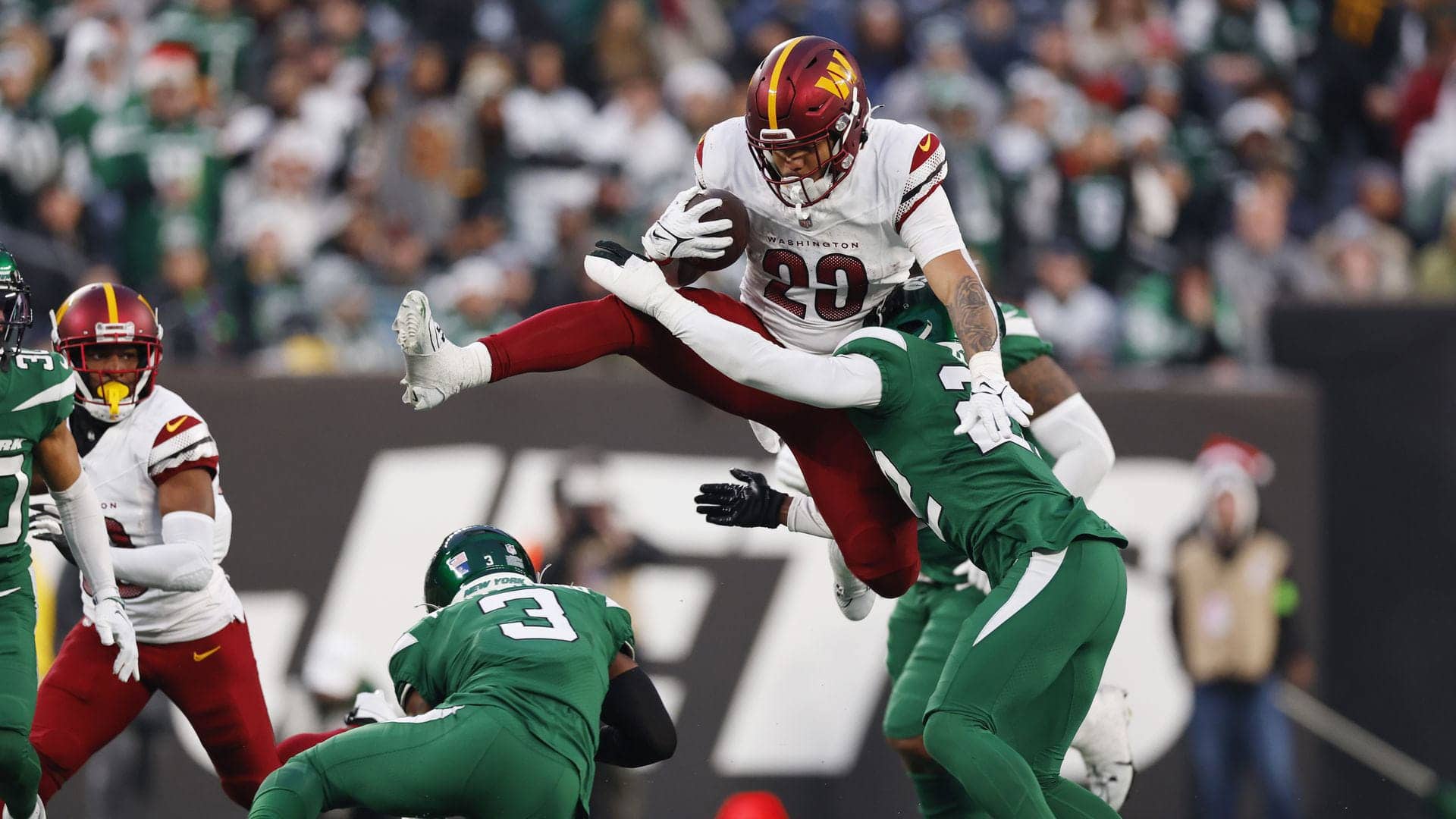 Football player in a white uniform leaps over an opponent while holding the ball during a game against a team in green.