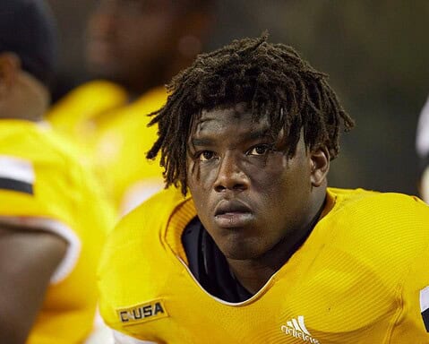 A football player wearing a yellow jersey with the text "CUSA," looking focused while on the sidelines.