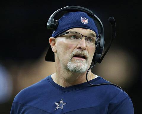 Coach in a blue NFL team shirt and a headset looks on during a game.