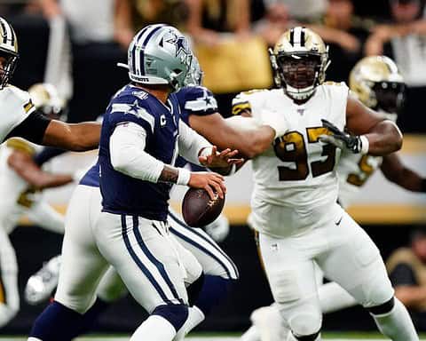 Football player in blue jersey evades two players in white jerseys on field during a game.