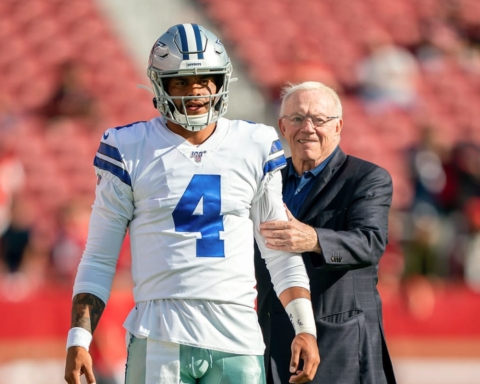 A man wearing a Dallas Cowboys football uniform stands next to an older man in a suit on a football field.