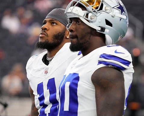 Two football players wearing white jerseys with dark blue numbers stand on the field, looking up.