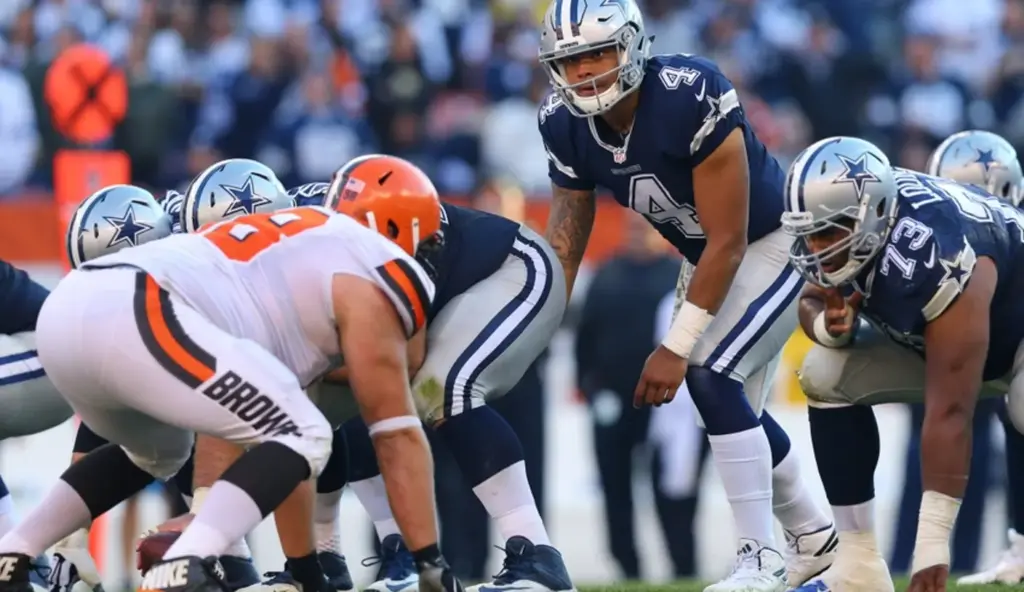 Football players from opposing teams line up for a play; a player in a blue uniform appears ready to receive the snap.